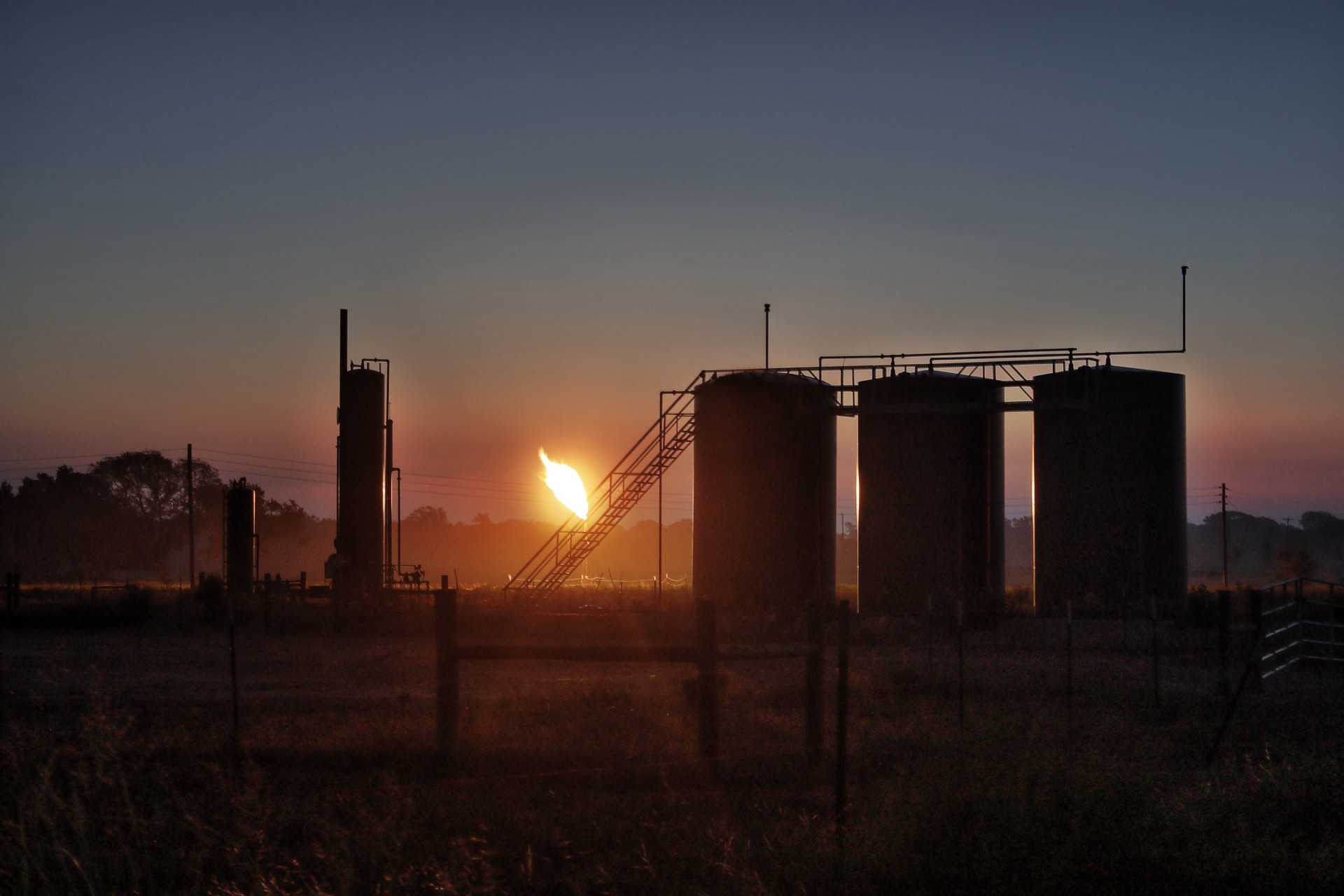 Wellsite storage tanks and a gas flare silhouetted against a setting sun in open prairie country. United States onshore production.